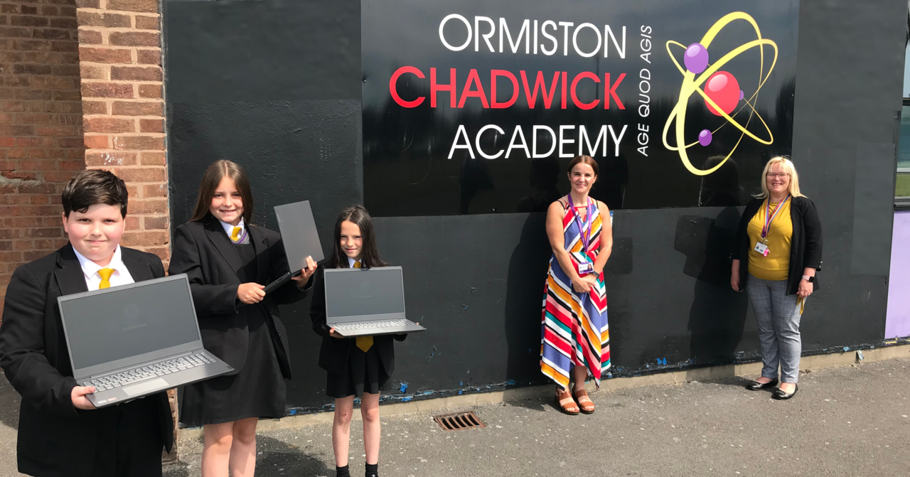 Three students with laptops stood in front of a school sign with two ladies, one from school and one from Halton Housing.